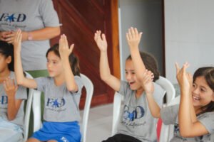 4 children sitting in white chairs in a circle worshipping with their hands in the air